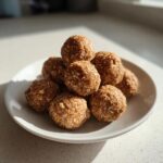 A stack of homemade No Bake Banana Cinnamon Energy Bites coated in oats, sitting on a white plate in bright sunlight.