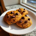 Three soft Pumpkin Chocolate Chip Cookies topped with whole chocolate chips, resting on a white plate near a sunny window.